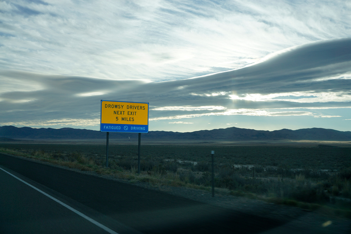 A bright yellow sign cautions drivers to exit if feeling drowsy on a stretch of highway in Nevada. The landscape is desolate and barren with a chain of mountains visible in the distance. The sun is low on the horizon hidden behind clouds.